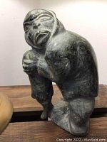 Front and side view of dark grey carved stone sculpture of a kneeling human figure with expressive face showing signs of wear.