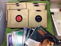Shows vinyl records in paper sleeves laid out flat on a green cloth surface, includes some visible labels and paper sleeves with wear signs.