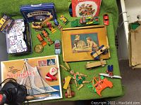 Photo showing collection of vintage toys laid on green fabric surface including domino sets, wooden pull toys, metal toy cars, framed board game and model kit box.