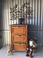 Wooden storage cabinet with three wicker front drawers, decorative metal wall art on top, ceramic vase and wooden bust sculptures placed around it, ornate clock in front