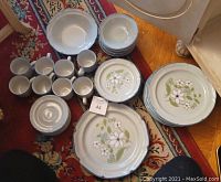 Photo shows the entire dining set arranged on a patterned rug with one large serving plate, dinner plates, salad plates, bowls, cups, and coffee plates.