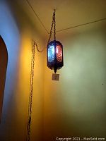 Photo of vintage hanging wall chandelier with colored glass panels lit up and hanging from the ceiling by a chain, anchored to a side wall with an additional chain link.