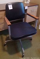Photo of black office chair with wooden armrests on metal base with wheels near filing cabinet and desk