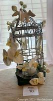 Silver birdcage styled decorative candle holder featuring two white glass birds perched on top and white artificial flowers wrapped around the frame and base. Candle inside the cage.