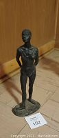 Full view of a decorative bronze-like figure statue of a person standing with arms behind back on an oval base.