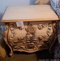 Photo of a wooden end table or night stand with three drawers showing carved floral and ribbon designs on front and sides, with cabriole legs.