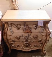 Front view of wooden end table with three drawers, detailed carved floral and ribbon patterns on drawer fronts and sides, light wood finish.
