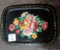 Top-down photo showing a black rectangular serving platter with a hand-painted floral design and a gold swirl border on a dark wooden floor.