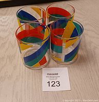 Four clear drinking glasses arranged in a tight group on a light-colored surface. Each glass decorated with overlapping colorful geometric bands in red, blue, yellow, and green.