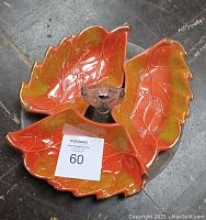 Three orange glass decorative dishes shaped like leaves placed on a round metal server tray on a stone floor surface.