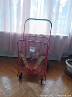 Front view of the red foldable metal wire grocery shopping cart with black handle grip and wheels, positioned indoors near window with curtains.