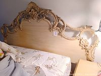 Photo of ornate beige carved wooden headboard attached to bedframe, showing floral and scroll details.