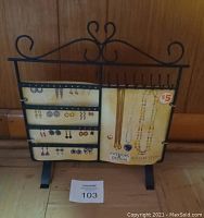 Photo of black metal jewelry holder with decorative top scroll, earring mesh panels on left and hooks on right holding two display cards with necklace and earring images.