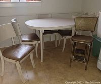 White oval kitchen table with four cream-colored chairs and one tall gold step stool chair arranged in a room on tiled floor.