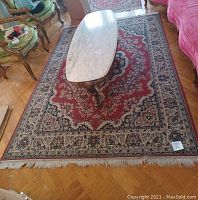 Full shot of rectangular living room rug under marble coffee table, showing red, cream, blue, and green floral traditional pattern with fringed edges.