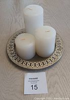 Round metal candle plate with a decorative pattern on the edge holding three white pillar candles of varying heights placed on a wooden surface.