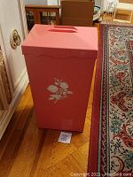 Photo of a pink plastic laundry basket with floral pattern standing on the floor near a carpet and furniture.