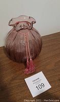 Photo of a purple tinted ribbed glass vase with a ruffled flared top and pink tassel tied around the neck, placed on a wooden surface.