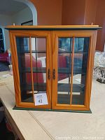 Small wooden trinket cabinet showing front face with two glass-paneled doors and metal handles. The cabinet is painted green on the outer edges with natural wood tone borders and black shelves inside.