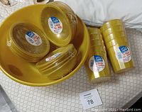 Ten yellow plastic cups, twelve regular size yellow plastic bowls, and one large yellow plastic bowl stacked on a bed.