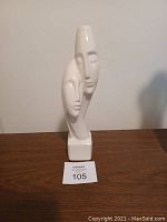 White glass decorative piece showing two overlapping stylized faces viewed from the front, placed on a wooden surface against a plain wall.