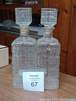 Photo showing two clear glass liquor decanters with matching square stoppers and raised geometric pattern.