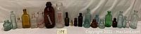 Full collection of 18 antique and vintage collectible bottles in various colors and shapes, arranged in a row.