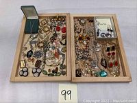 Overview of two wooden trays holding a wide assortment of costume jewelry including brooches, necklaces, and earrings arranged loosely