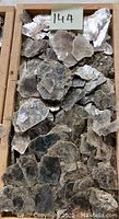 Full view of mica mineral pieces neatly stacked in a wooden tray, close-up shows typical thin, shiny flaky sheets in gray and brown hues.