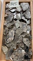 Top-down image of a wooden tray filled with multiple pieces of flaky mica mineral rocks, grayish and brownish in color, with a label '145' visible.