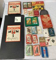 Top view of two Monopoly board games, various playing card decks, and boxed Lucky Clown pinball game arranged on a surface.