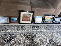 Six framed art pieces displayed on carpet in front of couch, showing various sizes and subject matter including landscapes and text.