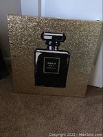 Front view of square canvas art showing black perfume bottle silhouette with 'Paris No 5 Eau de Parfum' label on densely gold glittered background.