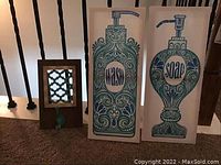Photo showing two tall canvas prints depicting decorative soap dispensers, and the wooden wall hook with a single metal hook on the carpeted floor near stair railing.