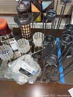 Two photos showing three silver metal multi-height candle holders, a decorative lantern candle holder with diamond pattern, two clear crystal glass tea candle holders, a pack of 50 white tealight candles, and a large orange candle on a glass table.