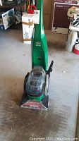 Front view of green Bissell upright carpet cleaner with handle and transparent cleaning head on concrete floor.
