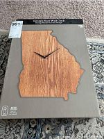 Photo of wall clock in original box showing the clock face shaped like Georgia state, wood grain finish, black clock hands, and box labeling.