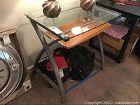 Front perspective of the glass and metal computer desk showing the clear glass top, metal frame with curved 'X' legs, and wooden pull-out keyboard tray. Two decorative lamps on top are excluded from the lot.