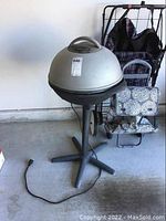 Full view of George Foreman electric grill with lid closed, standing on four-legged base with power cord visible on concrete floor alongside folded chairs.
