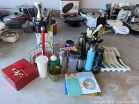Photo showing assortment of kitchen items including cookware, utensils, drink containers, and cook books arranged on a concrete surface.
