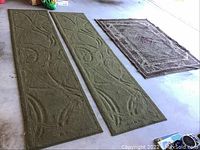 Three rugs laid on garage floor. Two long green runners with abstract raised pattern and one smaller woven multicolored rug.