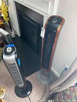Two tower fan heaters, one with wood grain finish and one silver with black, shown side by side near a fireplace.