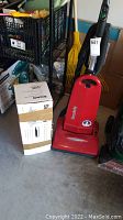 Red Simplicity upright vacuum cleaner standing on concrete floor near other garage items. Vacuum has a sticker on the front and black handle with controls visible.