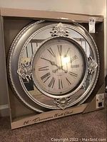 Clock in original box, showing front view of silver plastic framed clock with mirror accents and Roman numeral face.