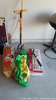 Photo showing various yard tools arranged on concrete floor including a metal rake, hand tiller, manual hedge clippers, work light, bags of plant food and soil, and boxes.