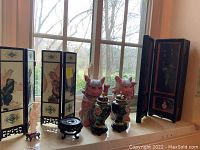 Overview of the Asian decor lot on a windowsill with natural light showing miniature silk screens, ceramic cats, urns, geisha figurine, and display stand.