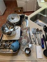 Overview of all items on a kitchen counter including cookpot, lids, measuring cups, knives, utensils, cutting board