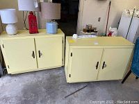 Pair of buttercream yellow painted wooden cabinets with two doors each, metal handles, topped with lamps and red decorative piece, set in garage floor environment.