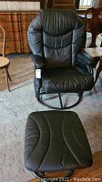 Black pleather reclining chair with matching ottoman set on carpet indoors, armrests and backrest padded, circular metal base visible.