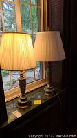 Pair of green and gold gilded table lamps with cream pleated shades on black marble surface by window.